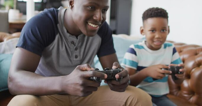Video of happy african american father and son sitting on sofa and playing video games