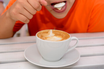 A cup of fragrant cappuccino close-up. In the background, a boy scoops up coffee foam with a spoon.