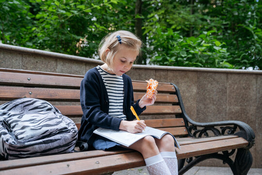 The girl is doing her homework and eating a sandwich in the park.