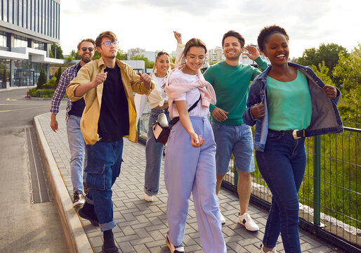 Group of multiracial friends are having fun walking along sidewalk on city street. Cool and stylish young people hang out together walking around city on summer day. Friendship and lifestyle concept.
