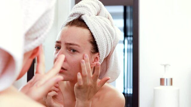 Worried young woman looking in mirror after shower, dissatisfied with skin condition, touching examining wrinkles on her face. Thinking of professional skincare products or cosmetologist meeting. 4k