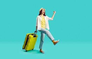 Happy woman with yellow suitcase going on summer holiday. Smiling overjoyed young girl wearing jeans, shirt and panama hat walking with her travel bag on bright turquoise background. Vacation concept - obrazy, fototapety, plakaty