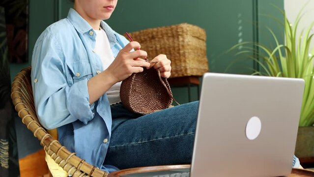 Young woman is engaged in crocheting, knitting for sale, learning watching tutorial online on video lessons sitting in living room.Creation of handmade basket accessory while watching movie on laptop