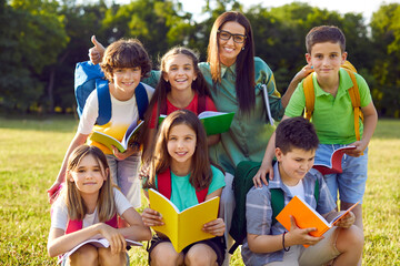 Outdoor portrait of happy modern Caucasian school children together with their teacher. Beautiful young woman and her elementary students posing for group photo in the park during fun lesson outside
