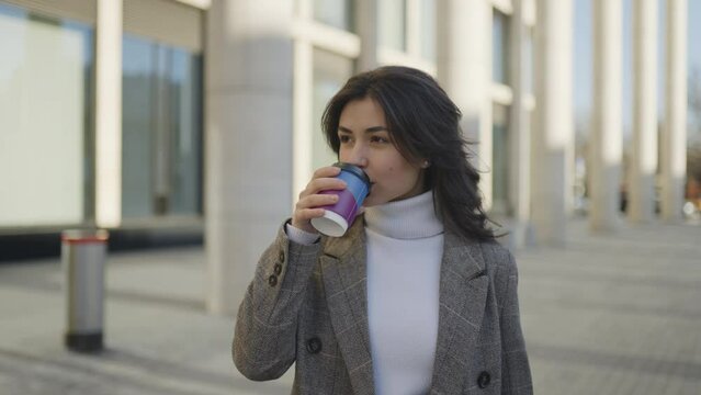 A young woman with dark hair and in a coat walks and drinks coffee, she enjoys and smiles. Against the backdrop of office buildings. Slow motion 4k footage