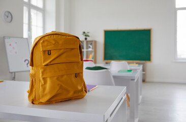 Fototapeta School classroom. New school bag on a student's desk in the classroom. Big yellow canvas backpack placed on the table in a large modern schoolroom with a chalkboard. Back to school concept obraz