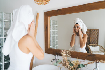 Young beautiful female with towel on head smiling to mirror reflection  standing in the bathroom at...
