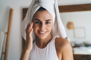 Young smiling female with towel on head puts moisturizing cream on face while standing at home...
