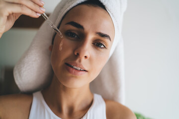 Young female with towel on head puts moisturizing serum on face while standing at bathroom. 30...