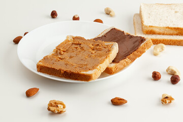 Square bread for toast with chocolate spread and peanut butter on a plate. Nuts, slices of bread and a white plate with sandwiches on a white table.