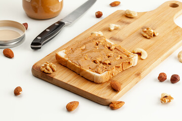 Square bread for toast with peanut butter on a wooden board. Nuts, a knife, a jar of peanut butter, and a wooden cutting board with a sandwich on a white table.