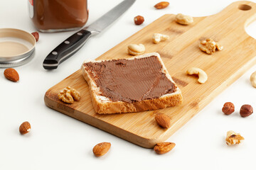 Square bread for toast with chocolate spread on a wooden board. Nuts, a knife, a jar of chocolate paste, and a wooden cutting board with a sandwich on a white table.