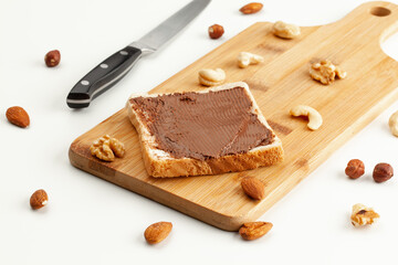 Square bread for toast with chocolate spread on a wooden board. Nuts, a knife, and a wooden cutting board with a sandwich on a white table.