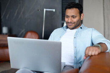 Smiling indian man spends leisure time in network, he lying down on the comfortable sofa with a laptop. Mixed-race guy making online shopping, web surfing, working remotely