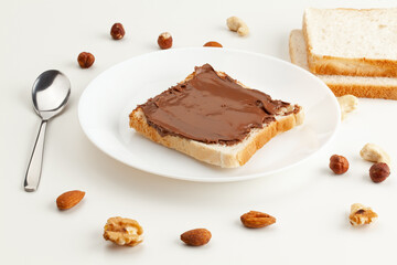Square bread for toast with chocolate spread on a plate. Nuts, a spoon, slices of bread and a white plate with a sandwich on a white table.