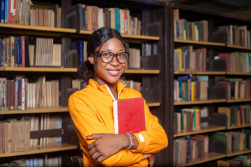Cheerful female international student standing beside bookshelf in the library