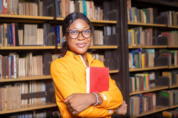 Cheerful female international student standing beside bookshelf in the library