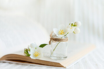 Close-up of a bouquet of white flowers in a blurred glass vase on an open book on a white knitted background. Slow life concept.