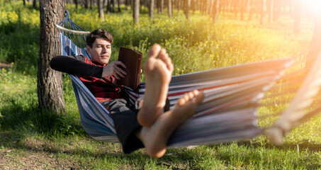 young blond man lying on a hammock with bare feet in a forest, while reading a book. relaxation and...