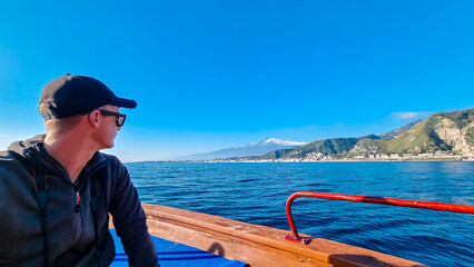 Man on touristic boat tour with panoramic view from open sea on snow capped volcano Mount Etna and...