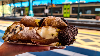 Person holding homemade Sicilian Cannoli with ricotta filling garnished with pistachio, sugar powder. Panoramic view of snow capped Mount Etna volcano on sunny day. Taormina, Sicily, Italy, Europe, EU