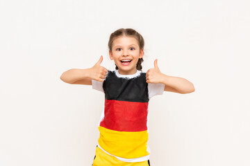 A little girl with a German flag on a T-shirt, with long hair in pigtails, gives a thumbs up and smiles broadly on a white isolated background. Language courses.