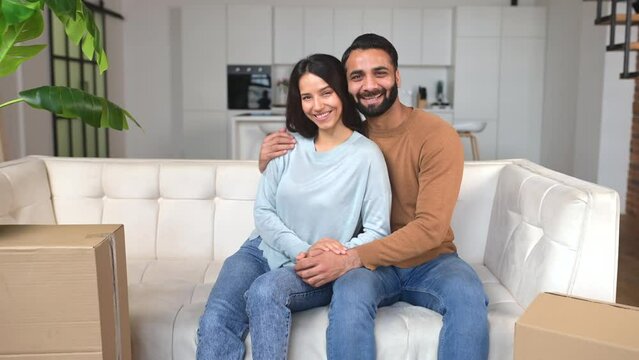 Happy smiling Indian couple in love sitting on the sofa in surrounded with cardboard boxes, looking at the camera and smiling, ethnic young girlfriend and boyfriend relocated, moved in new flat