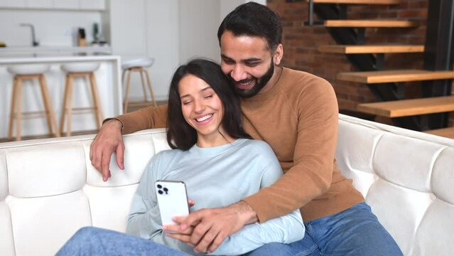 Positive Indian couple in love spends time together at home, using smartphone for web surfing, smiling pakistani guy and woman looking at phone screen together, scrolling news, or choosing smth online
