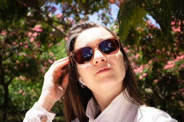 Young beautiful girl in stylish sunglasses against Chinese acacia pink blossoming flowers in spring garden. Portrait of young woman in blooming spring, summer park. Romantic vibe. Female and nature.