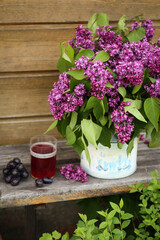 Summer atmosphere, garden. A bouquet of lilacs in a white vase on a wooden bench. A glass of compote, grapes. Rustic. Background image, copy space
