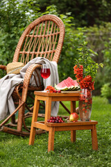 Summer garden. Rocking chair, straw hat, green trees. Bouquet with red flowers, fresh watermelon, pear and cherry on the table, stool. A glass of red wine. Summer atmosphere. Background image
