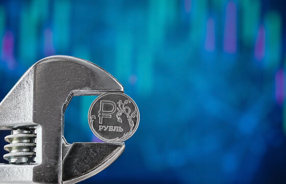 A coin with the symbol of the Russian ruble on a blurred blue background with a graph of falling quotes is squeezed with a wrench. News about the default and the collapse of the economy. Free text