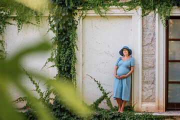 Portrait of middle-aged pregnant woman in denim blue dress, hat. The future mother stands near antique ruins, grunge background with green ivy. Travel during pregnancy. Maternity and pregnancy concept