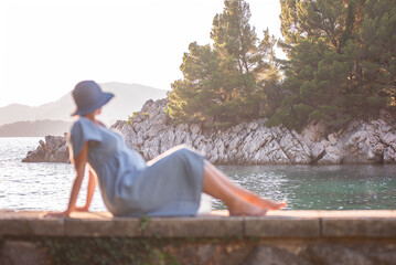 Defocused tender pregnant woman in denim dress, blue hat sits by the sea in the sun against the backdrop of green island. Traveling in pregnancy to Montenegro on the Royal Island. Mothers care, rest