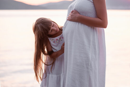 Close-up portrait of a little girl hugging pregnant mother. A daughter in a white sundress strokes a belly to a middle-aged woman by the sea at sunset. Family travel. The concept of happy motherhood