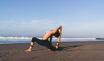 Attractive Caucasian woman in tracksuit practicing yoga exercises feeling balance and peace, strong fit girl with perfect figure standing in asana during morning training at seashore coastline