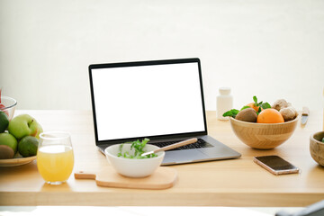 Laptop computer with white screen on wooden table in the kitchen with fruits and vegatables, copy space. Food blogger concept