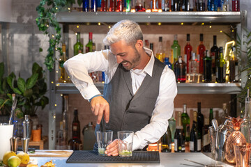 Happy and handsome barman making mojito cocktail in night club preparing expert drinks on bar counter smiling.