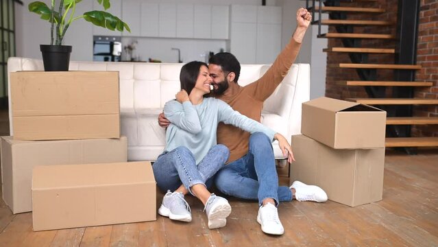 Happy Indian newlyweds moved in new apartment, sitting on the floor among cardboard boxes and celebrating moving day, raising hands up, multi ethnic couple in love rent a house together, relocated