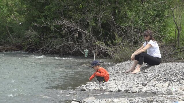 Little boy playing with a rod on mountain river bank near his mother