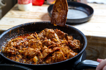 Chicken pieces fried in sauce in spices in a deep frying pan with a wooden spatula.