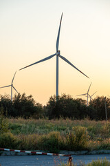 Large windmill against backdrop of rising sun in yellow and white tones behind green forest and...