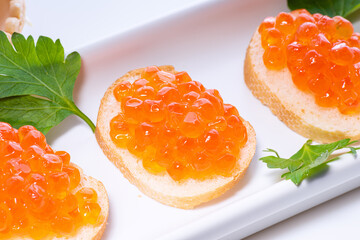Red caviar decorated with green parsley on small butter buns on white rectangular plate on white background.