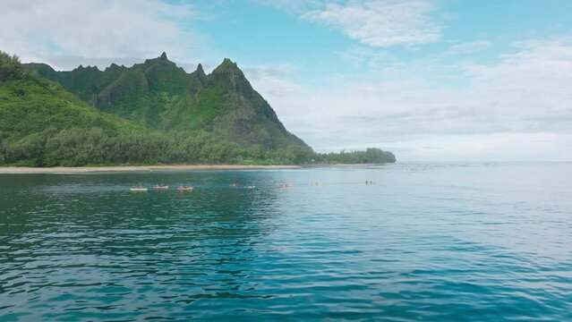 Cinematic aerial view group of tourists in colorful kayaks rowing in blue open waters of Pacific ocean. Kayakers on green mountain motion background, Napali park coast kayak tour, Kauai island Hawaii
