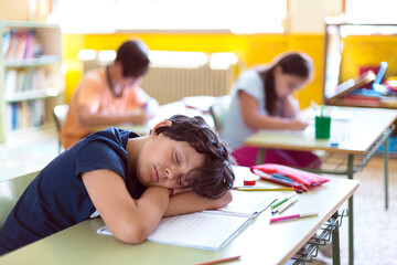 Caucasian child sleeping in class while his classmates are doing homework.