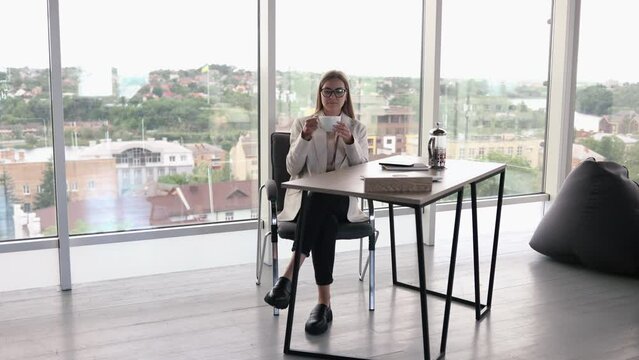 Pretty young woman in glasses working in a bright office and drinking tea at a laptop. Attractive long haired lady with glasses talking on the phone