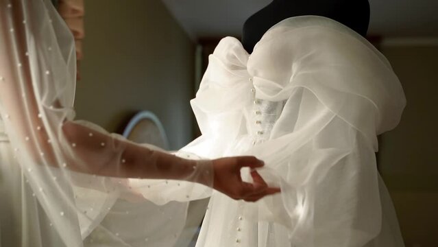 Bride enters the room in long bridal veil, strokes and touches her wedding dress with her hands. Woman admires her white dress.