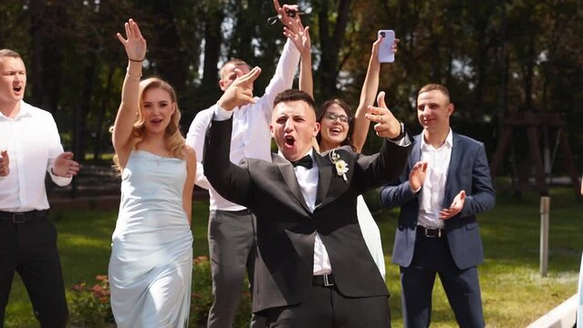 Groom flexing having fun with groomsmen on wedding day. Happy classy man in suit, bow tie and boutonniere hanging out partying with friends and shouts at camera before ceremony.
