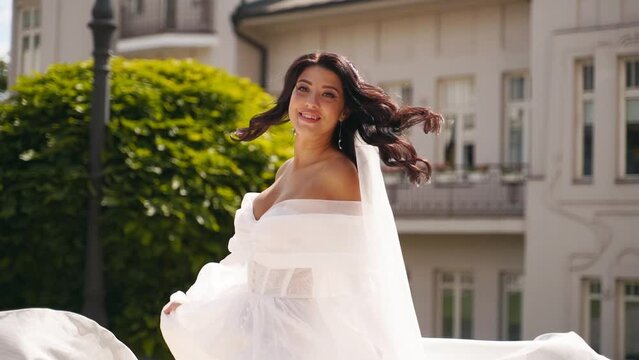 Pretty bride in long airy veil, elegant white dress spins with fabric fluttering in air on wedding day in luxury palace garden. Playful attractive woman posing on camera swirling in fabulous gown.