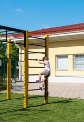 A 6-year-old Caucasian girl goes in for sports, climbs on a gymnastic wall on an open sports ground.